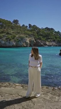 Beyaz elbiseli bir kadın, Akdeniz manzarasını çevreleyen yemyeşil yemyeşil yemyeşil yemyeşil yemyeşil, Mallorca 'daki Turkuaz suları ve kayalık uçurumlarda güneşli bir günün tadını çıkarıyor..