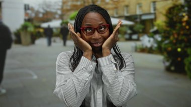 Kıvırcık saçlı, gözlüklü bir kadın şehir arka planında neşeli ifadeler takınıyor..