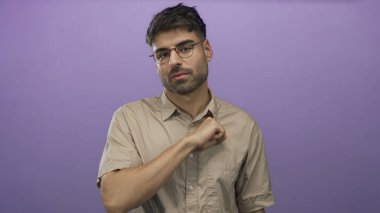 Gözlüklü ve bej gömlekli İspanyol genç adam mor stüdyoda göğsünü yumrukluyor, samimiyet..