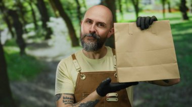 Sakallı ve eldivenli bir adam açık havada, yeşil bir parkta tulum giymiş, kendine güveni ve cazibesi olan yumuşak gülüşü ve kapalı gözleri olan bir kağıt torba tutuyor..