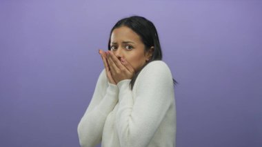 İzole edilmiş mor arka plan üzerindeki genç İspanyol kadın, elleri yüzü ve endişeli ifadesiyle korkusunu ifade ediyor, beyaz bir kazak giyiyor ve kaygı gösteriyor..