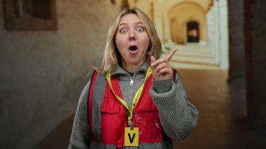 Sarışın bir kadın, eski bir üniversite avlusunda düşünceli bir şekilde poz vermeye gönüllü. Rozetli kırmızı bir yelek giyiyor, tarihi mimariyle çevrili..