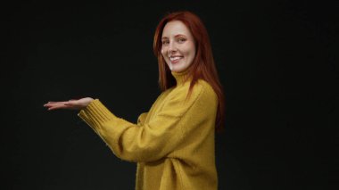 Kadın, siyah arka plana doğru elini uzatıyor sarı süveter giyiyor özgüvenli ve çekici görünüyor uzun kızıl saçlı izole stüdyo ortamında..