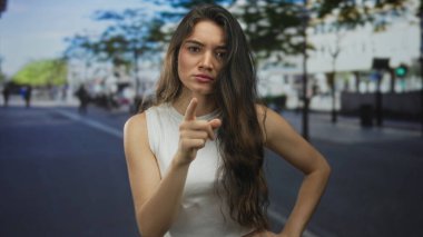 Yapraklı ağaçların altında güneşli şehir sokağında objeyi işaret eden Latin kökenli genç kadın..