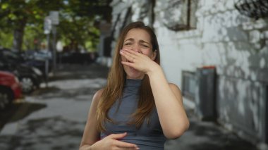 Uzun kahverengi saçlı, beyaz bir kadın, şehir caddesinde elleri kolsuz bluzuyla ağzını kapatıyor..