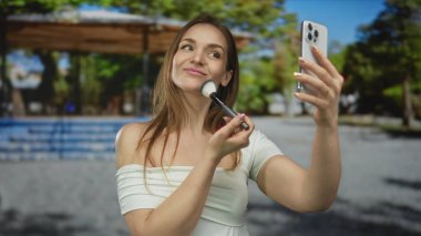 Akıllı telefon tutarken makyaj fırçası süren ve parkta gülümseyen bir kadın; güzellik özgüveni.