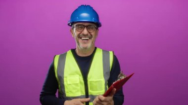 Siyah şapkalı ve güvenlik yeleği giymiş gülümseyen yetişkin bir adam elinde bir pano, izole edilmiş pembe bir arka planda duruyor.