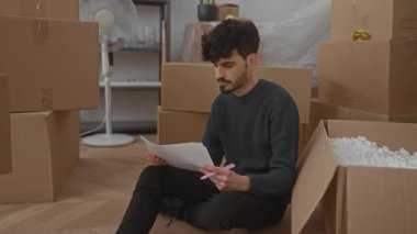 Oturma odasındaki kutularla çevrili yeni bir evin zemininde oturan genç adam dikkatle belgelere bakıyor..