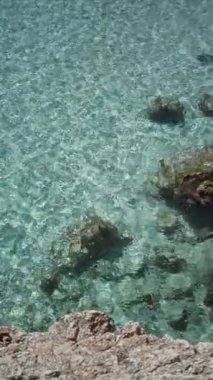 Berrak turkuaz su, parlak güneş ışığı altında kayalık sahil şeridinde yavaşça tur atar..