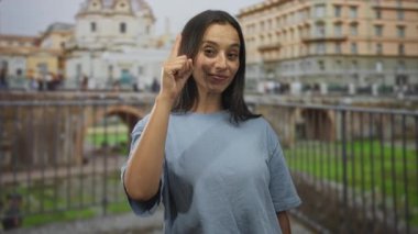 Kadın işaret parmağıyla dışarıdaki bir arkeolojik sitede yıkılmış bir Roma binasını işaret ediyor, gülümsüyor ve kameraya bakıyor;.