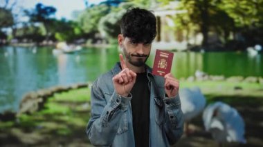 Yeşil ağaçlar ve suyla dolu bir parkta İspanyol pasaportu taşıyan bir adam kot ceket giyiyor, geziyor ve açık havada kimlik gösteriyor..