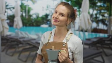 Önlüklü bir kadın güneşli bir günde arka planda palmiye ağaçları ve şezlonglarla havuz başında kahve makinesini tutuyor.