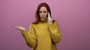 Sarı kazaklı, kırmızı saçlı bir kadın telefonda soyutlanmış pembe arka plana karşı konuşurken hayal kırıklığına uğramış görünüyor. El hareketleriyle ve yüz ifadeleriyle duygularını gösteriyor..