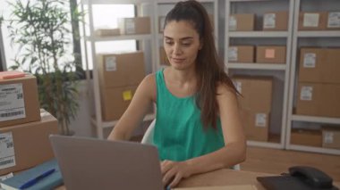 Dizüstü bilgisayarda yazan kadın, ofisindeki nakliye paketlerinin arasında elleri klavyede; küçük işletme gururu.