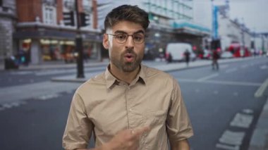 Gözlüklü ve bej tişörtlü bir adam yoğun bir şehir caddesinde gündüz vakti parmağıyla işaret ediyor; onay.