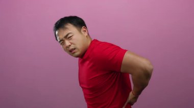 Kırmızı gömlekli, sırt ağrısı olan genç bir adam canlı pembe bir duvara karşı izole edilmiş, yüzünü buruştururken, sağlık sorunlarını ve vücut dilini vurgularken rahatsızlığını ifade ediyor..