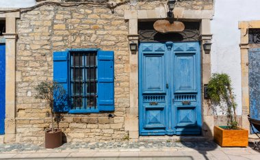 Güneş ışığı, canlı mavi kapı ve pencere panjurları olan, geleneksel mimariyi geliştiren taş bir evin resimsel görüntüsünü aydınlatıyor.