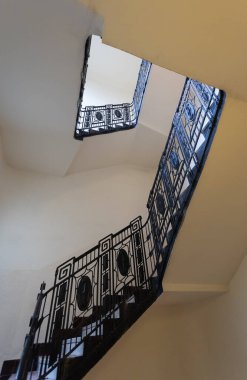 Dekoratif siyah metal parmaklıklı zarif bir merdiven manzarası, bir binanın üst katlarına doğru yükseliyor.