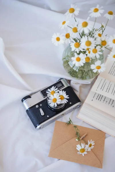 Chamomile flowers in vase, vintage camera and book on white fabric.