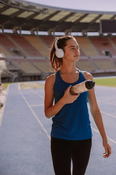 An attractive female sportsperson is walking at the stadium with bottle in her hands. She is taking a break from running