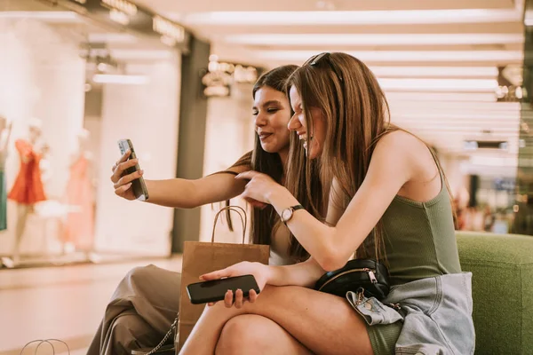 两个漂亮的女孩坐在购物中心的沙发上 一边看着电话 一边笑 — 图库照片