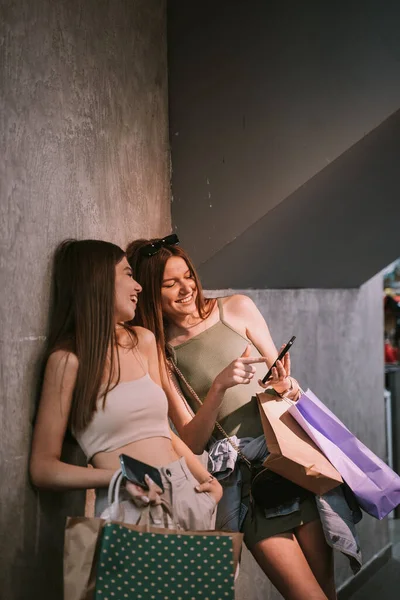 Two young beautiful sisters laughing at their phone whilst holding bags with clothes inside a shopping mall