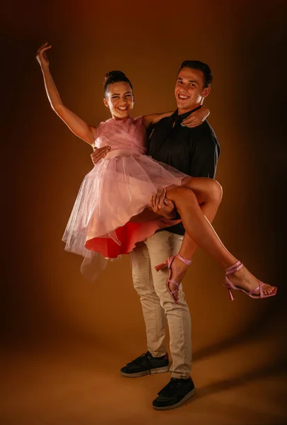 A smiley couple in studio. Boy took his girlfriend in his arms. She is wearing dress and high heels and he is in shirt, pants and sneakers