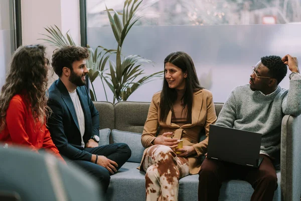 Diverse positive group of people taking their first coffee at work early in the morning. They are sitting on a gray sofa and having fun conversaiton