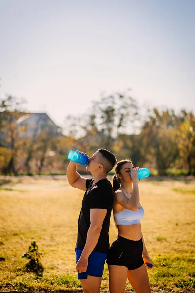 Sexy couple standing back to back and drinking water after workout in the park on a sunny day