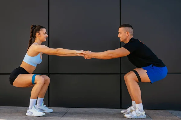 An attractive sports couple with resistance bands around their legs, holding their hands and doing squats