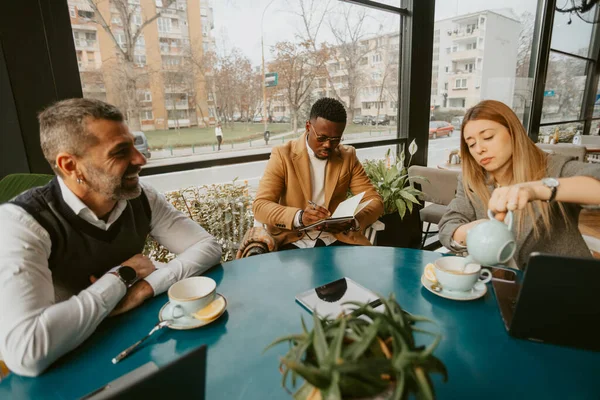 Blonde business woman pouring tea in her blue cup while the business man in the middle is writing and the third is smiling and looking at the her