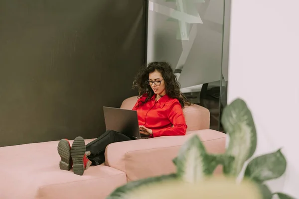Full length photo of gorgeous curly haired business woman dressed in red shirt, black pants and red boots lying on a pink sofa at modern office working on her lap top