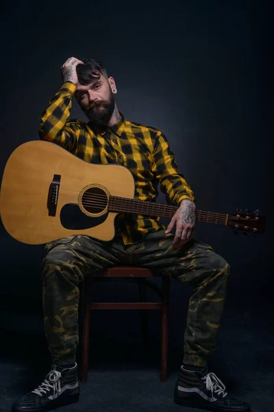Man with tattoos and beard is hoding a guitar and is wearing a tartan yellow black shirt
