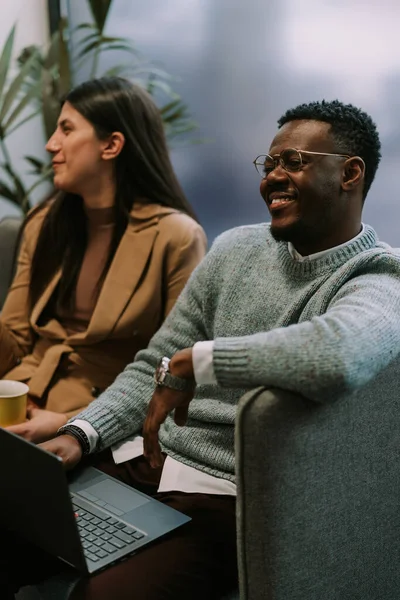 Good looking black male person smiling while looking at his colleagues having fun at the office. He has a lap top on his lap