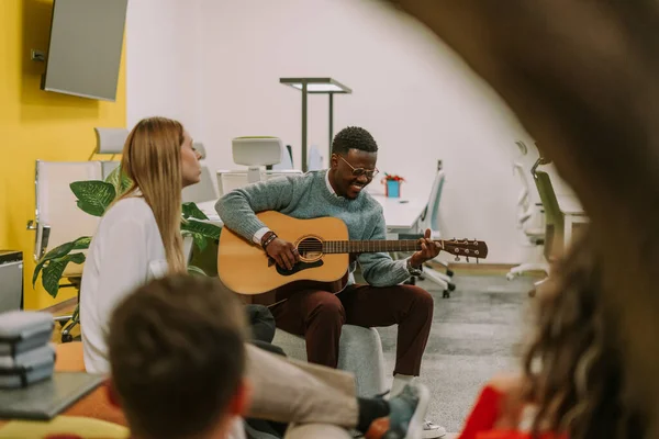Good looking black man smiling and playing guitar for his colleagues. His white blonde female coworker is singing