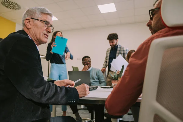 Stock image Close up of two older business men talking while sitting with their younger colleagues at the co working space