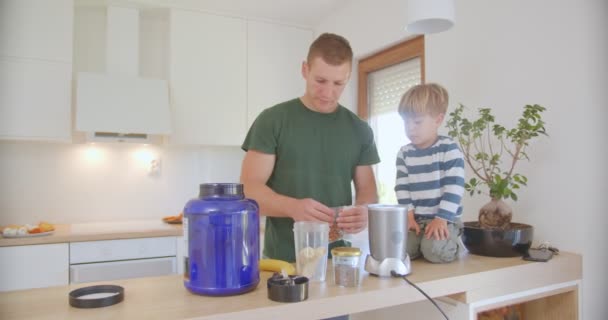 Far Och Son Lägger Nötter Banansmoothien Medan Sonen Sitter Köksbänken — Stockvideo
