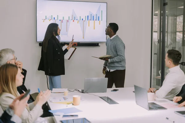 A stylish multiracial couple explaining financial charts details to their colleagues on presentation monitor