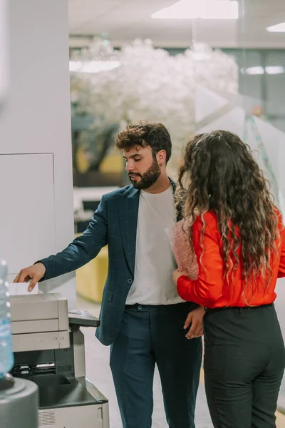 A good looking businessman printing documents and giving new instructions to his curly haired secretary standing next to him