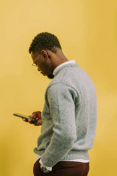 一位长相漂亮 时尚的黑人男性商人站在黄色背景下 一边在办公室休息一边检查自己的手机 — 图库照片