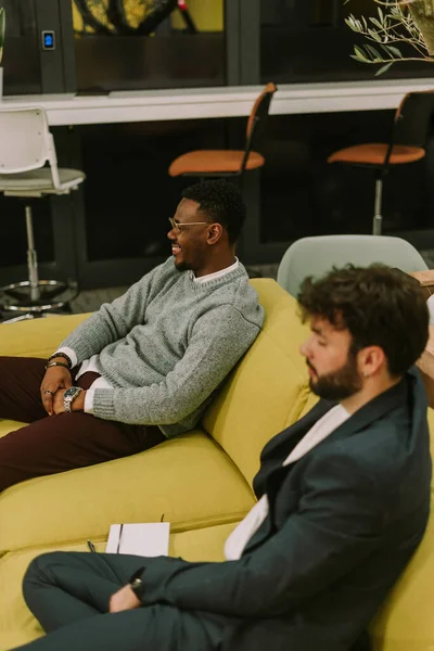 A tired business person felt asleep while taking a break and sitting on yellow sofa at modern office. His black male is looking at his colleagues having fun and smiling