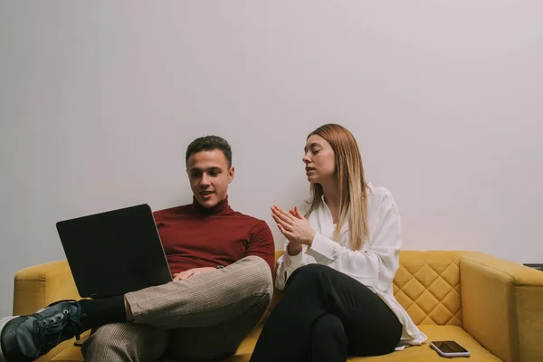 Good looking male person showing something on the lap top to his female colleague. They are taking a break from work and sitting on yellow sofa at modern co working space
