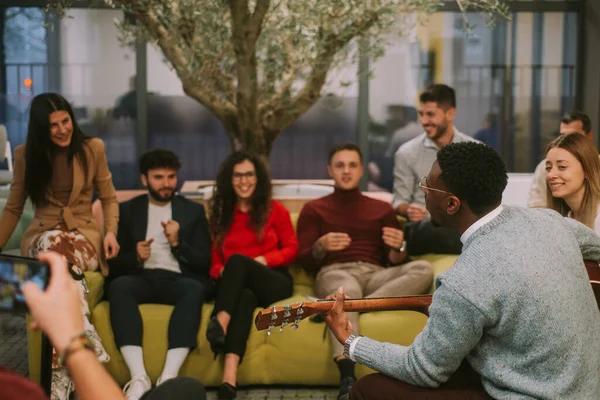 Group of young people enjoying listening to their black male colleague playing guitar and singing for them at the office after rough day at work