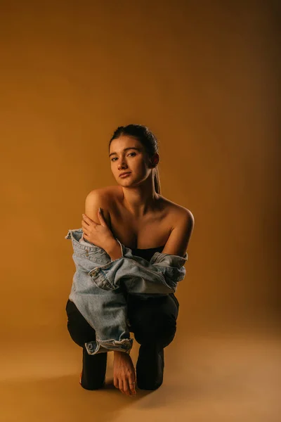 Fashion shoot in studio. Girl crouching and posing in black pants and blue jacket. She is touching her right shoulder with the left arm