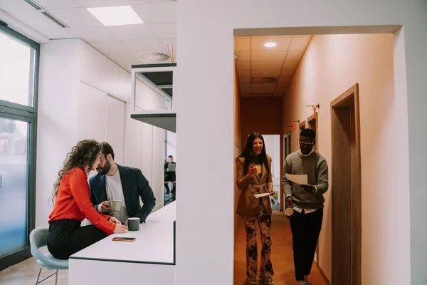 オフィスキッチンでコーヒータイムを楽しみながらイチャイチャする素敵なビジネスカップル 彼らの多人種のカップルの同僚は壁の反対側で楽しい会話をしている — ストック写真