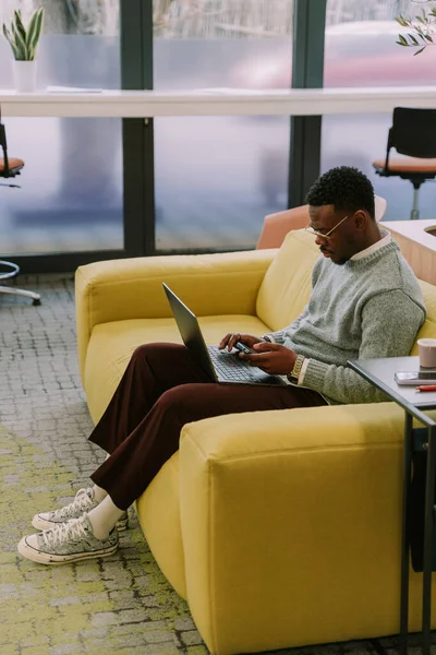 Good looking black male person pretending that he is working on his lap top, but actually he is spending time on his phone