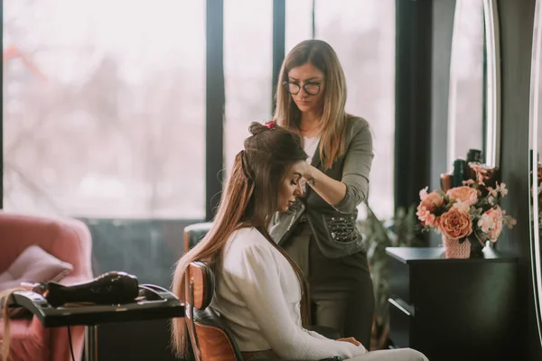 当她坐在美发厅时 金发美发师正在给她的顾客刷直棕色的头发 — 图库照片