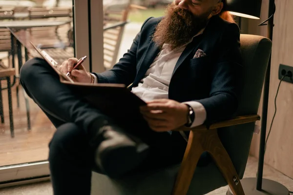 一个英俊的留着胡子的男人 双腿交叉地坐在咖啡店的椅子上 在记事本上写着什么 — 图库照片