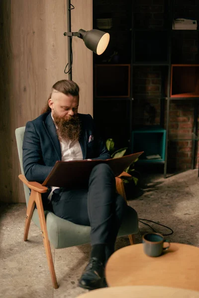 一个留着胡子的年轻人坐在咖啡厅里 两条腿交叉坐在笔记本上读书写字 — 图库照片