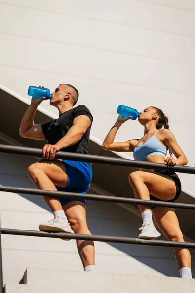 stock image Urban sporty couple drinking water after training on a sunny day. They are taking a break. Low angle view